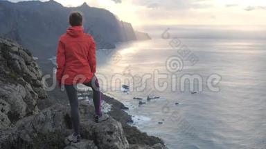 一位年轻女子<strong>站在山顶</strong>上，靠近海洋上的悬崖边缘，在特内里费岛上观察日落。 夫人在